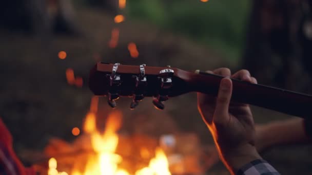 Close-up shot van mannelijke toeristen hand gitaar spelen tijdens avond op Camping met vuur verbranden in de achtergrond. Muzikale instrumenten, de natuur en de mensen concept. — Stockvideo
