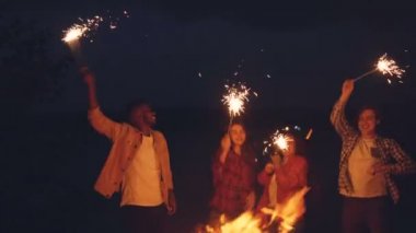 Heyecanlı gençler eğleniyor ve gülüyor zevk müzik, parti ve doğa orman yangında bengal ışıklar ile dans ediyor. Ağaçlar ve göl görülebilir.