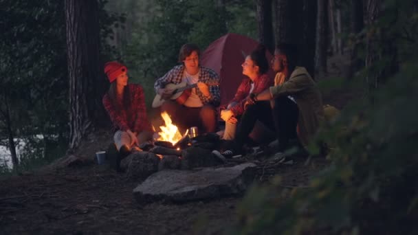 Los viajeros alegres están cantando canciones y tocando la guitarra sentados alrededor del fuego en el bosque por la noche y divirtiéndose con la hermosa naturaleza alrededor. Gente y concepto musical . — Vídeos de Stock