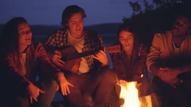 Homens e mulheres jovens estão cantando músicas para a guitarra descansando ao redor da fogueira e desfrutando de música e boa companhia na noite de verão. Árvores e lago são visíveis . — Vídeo de Stock