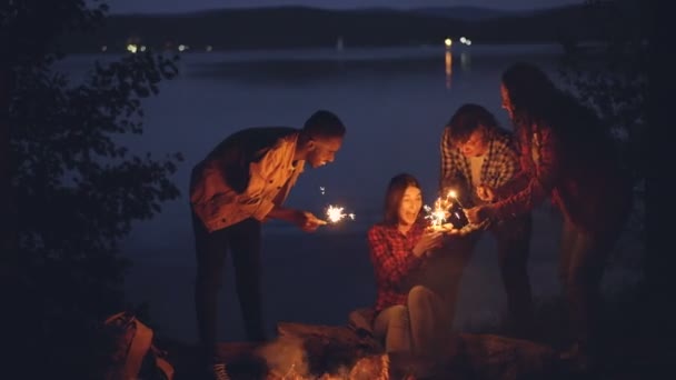 Amigos cariñosos turistas están felicitando a la chica en el cumpleaños trayendo pastel y quemando luces de bengala, la mujer está soplando velas, sosteniendo el brillo y gritando . — Vídeos de Stock