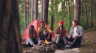 Neşeli kızlar ve çocuklar yürüyüşçü konuşuyor ve hikayelerini paylaşan ve having fun kamp ateşi yanında oturan gülüyor. Yeşil ağaçlar, modern çadır ve alev görülebilir.