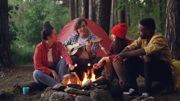 Les randonneurs heureux cuisinent la guimauve sur le feu et chantent des chansons tandis que le beau gars joue de la guitare lors d'une escapade en forêt. Concept nature, amusement et amitié . — Video