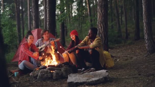 Расслабленные путешественники поют песни у костра в лесу, играют на гитаре и жарят зефир. Видны зеленые деревья и палатка . — стоковое видео