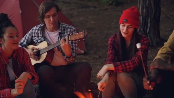 Friends at campsite are singing songs, laughing and cooking food on fire while happy smiling guy is playing the guitar. Camping, music and friendship concept. — Stock Video