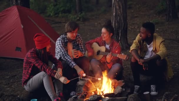 Jolie femme joue de la guitare pendant que ses amis chantent des chansons drôles et rient en faisant cuire la guimauve sur le feu et en applaudissant les mains. Musique, nature et concept amusant . — Video