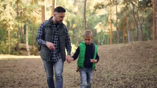 Adorabile ragazzo sta camminando nella foresta con il suo amorevole padre che gli tiene la mano, barbuto giovane sta parlando e gesticolando. Escursioni, famiglia e concetto di paternità . — Video Stock