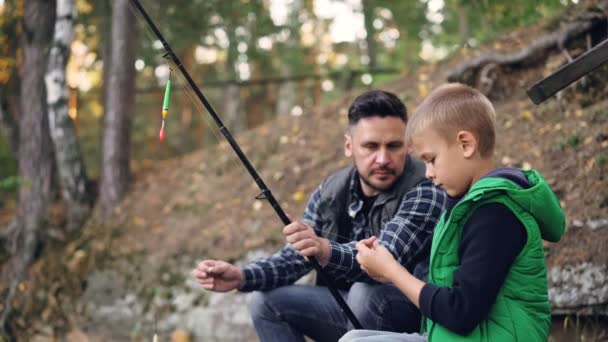 Padre e figlio pescano con le canne e parlano, l'uomo insegna al bambino ad usare l'attrezzatura nella giornata autunnale con gli alberi intorno. Famiglia, hobby e tempo libero . — Video Stock