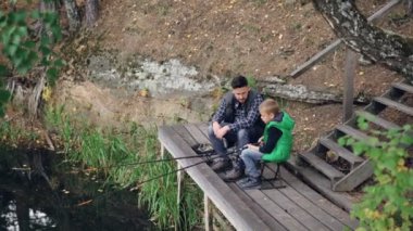 Küçük çocuk gölde ahşap iskele babasından ile sonbahar gününde balık tutuyor, çocuk babası onunla konuşurken sandalyede oturan rod tutuyor. Doğa ve insan kavramı.