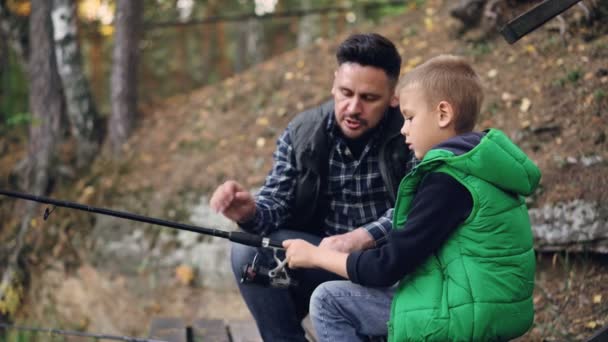 Adorable niño está pescando con papá en el estanque en el bosque sosteniendo la caña, mientras que su padre está hablando con él explicando cómo utilizar el equipo. Familia, afición y concepto de naturaleza . — Vídeos de Stock