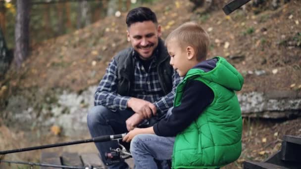 Бородатий молодий чоловік рибалить з милою дитиною в осінній день, хлопчик тримає палицю і розмовляє з батьком, навчаючись ловити рибу. Люди, обмін досвідом та концепція сім'ї . — стокове відео