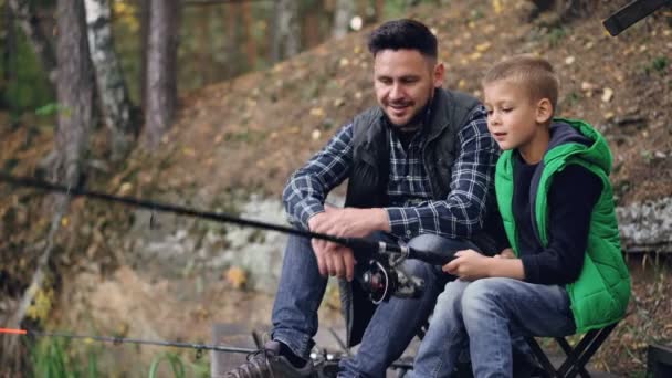 Красивый парень, любящий отца, учит своего сына ловить рыбу, сидя на берегу реки вместе, мальчик держит удочку и двигает ее, разговаривая с папой. Хобби, приключения и семейная концепция . — стоковое видео