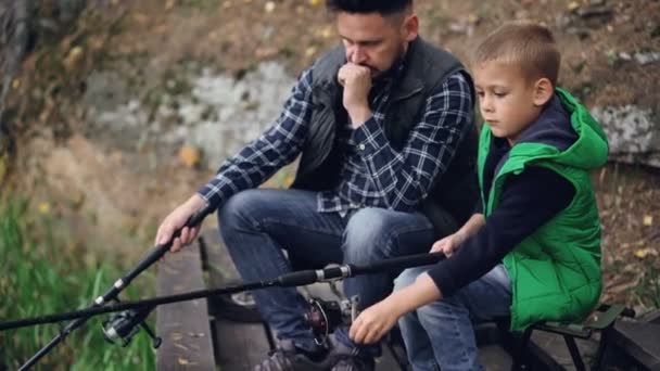 Allvarlig liten pojke är att fånga fisk med sin far skäggiga brunet sitter tillsammans och hålla fiskespön. Gemensamma hobby, generationer och familjen koncept. — Stockvideo