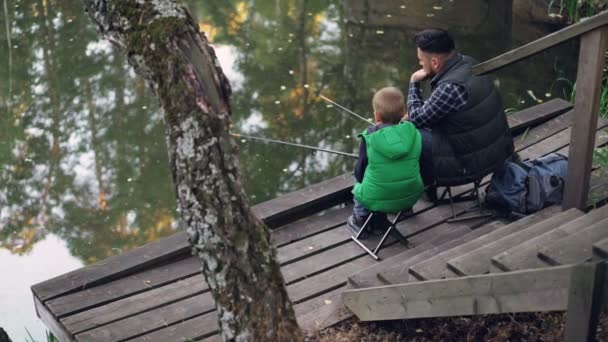 Vysoký úhel pohledu osoby otce a syna rybaření v rybníčku v lese sedí na židlích na dřevěném molu a držení tyče. Pojem rodina, hobby a příroda. — Stock video