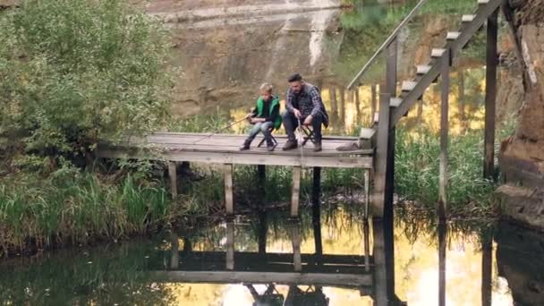 カジュアルな服でハンサムな男は棒と秋晴れの美しい池を見てウッドデッキの上に座って彼の子供と魚を捕る。自然とレクリエーションの概念. — ストック動画