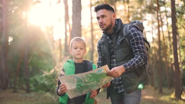 Mouvement lent du petit garçon tenant la carte debout dans la forêt pendant que son père parle et fait un geste enseignant à son fils l'orientation dans le bois. Nature, famille et concept de navigation . — Video