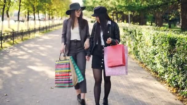 Twee aantrekkelijke jonge vrouwen lopen van winkels langs mooie straat uitvoering van papieren zakken met aankopen en praten over de mooie herfstdag. Winkels en concept. — Stockvideo