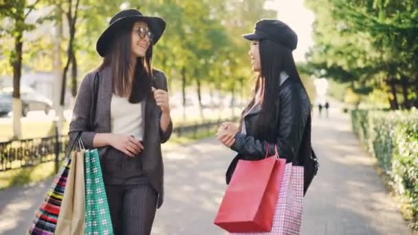 Giovani amiche felici stanno parlando, ridendo e abbracciandosi in piedi in strada con sacchetti di carta che condividono notizie. Shopping, amicizia e concetto di persone . — Video Stock