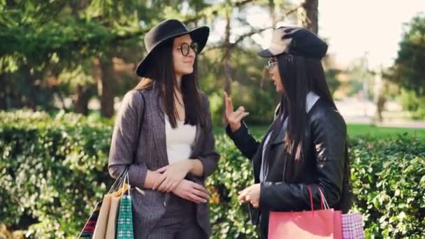 Jolie fille asiatique parle à son amie jolie brune debout dans la rue tenant des sacs à provisions et riant. Concept de personnes, de villes et de commerçants . — Video