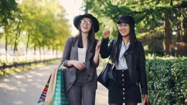 Tilt-down shot of happy female millennials shopaholics walking together with bright shopping bags and talking. Las mujeres están usando ropa y gafas de moda . — Vídeos de Stock
