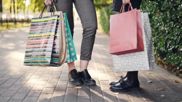 Baixo ângulo tiro de pernas femininas em pé na calçada segurando sacos de compras depois de feliz dia ocupado em lojas e shoppings. Pessoas, consumismo e conceito de lazer . — Vídeo de Stock