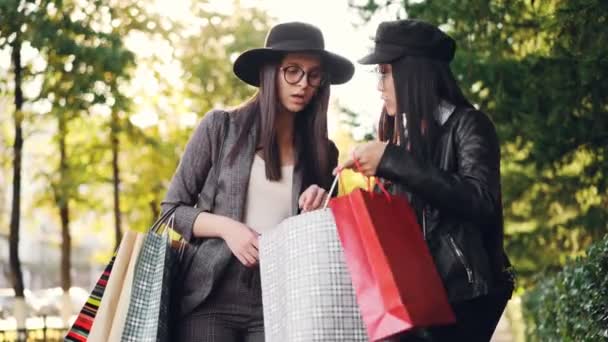 Возбужденная девушка смотрит на своих друзей покупки в сумках, задавая вопросы о ценах и коллекциях и выражая волнение о покупках стоя на улице в городе . — стоковое видео