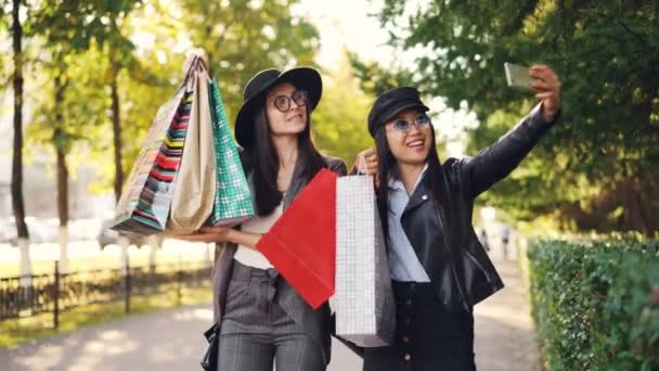 Ευτυχισμένος νεαρές γυναίκες φίλοι παίρνουν selfie με χάρτινες τσάντες στέκεται έξω στο πεζοδρόμιο και να ποζάρουν. Κορίτσια είναι χρησιμοποιώντας το smartphone και να φοράει ρούχα. — Αρχείο Βίντεο