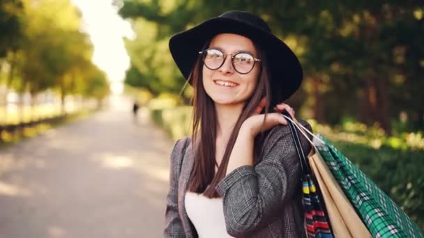 Retrato de la joven morena feliz mirando a la cámara y sonriendo sosteniendo bolsas de compras en la mano de pie al aire libre. Ciudad, pueblo y concepto de consumismo . — Vídeos de Stock