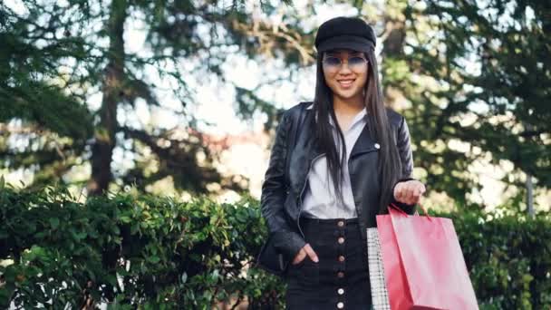 Porträt einer lächelnden asiatischen Shopaholic, die mit Einkaufstüten auf der Straße steht und in die Kamera blickt. Im Hintergrund grüne Bäume und Sträucher. — Stockvideo
