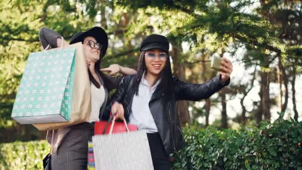 Chicas emocionadas están haciendo videollamadas en línea utilizando un dispositivo de retención de teléfonos inteligentes y mostrando bolsas de compras con compras. Comunicación, tecnología y concepto divertido . — Vídeos de Stock