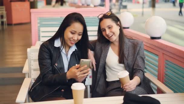 Bonitas amigas jóvenes están usando un teléfono inteligente moderno sentado en la mesa en la cafetería con café para llevar. Chicas felices están tocando la pantalla, viendo fotos y riendo . — Vídeo de stock