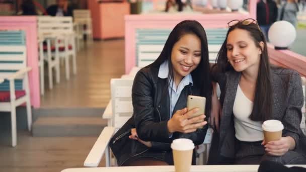 Les jeunes femmes gaies se détendent dans le café et utilisent un téléphone intelligent regardant l'écran et souriant tenant pour aller café. Concept jeunesse, technologie et boissons . — Video