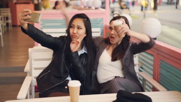 Meninas despreocupadas estão tomando selfie com tirar bebidas sentadas no café juntos e usando smartphone. Mulheres jovens estão posando, batendo óculos e mostrando gestos de mão . — Vídeo de Stock