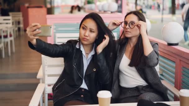 Hermosas mujeres jóvenes están tomando selfie en la cafetería sentado en la mesa y el uso de teléfono inteligente. Atractiva chica caucásica está usando gafas, su amigo asiático está sosteniendo gadget . — Vídeo de stock