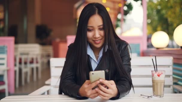 Sonriendo estudiante chica asiática está descansando en la cafetería al aire libre y el uso de la pantalla táctil del teléfono inteligente y ver. Redes sociales, tecnología moderna y concepto de ciudad . — Vídeos de Stock