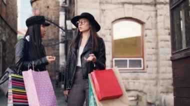 Şirin Asyalı kız alışveriş torbaları tutarak ve gülüyor sokakta onu beyaz arkadaş zarif esmer ayakta konuşuyor. Kişi, şehir ve shopaholics kavramı.