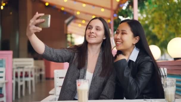 Alegre amigos femeninos están haciendo videollamada en línea con teléfono inteligente mirando a la pantalla y hablando sentado en la cafetería de la calle juntos. Concepto de comunicación y tecnología . — Vídeos de Stock