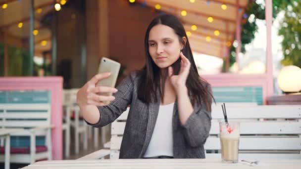 Hermosa chica con el pelo largo y oscuro se está tomando selfie y posando para la cámara del teléfono inteligente sentado en la cafetería de la calle solo. Estilo de vida moderno, la juventud y el concepto gadgets . — Vídeo de stock