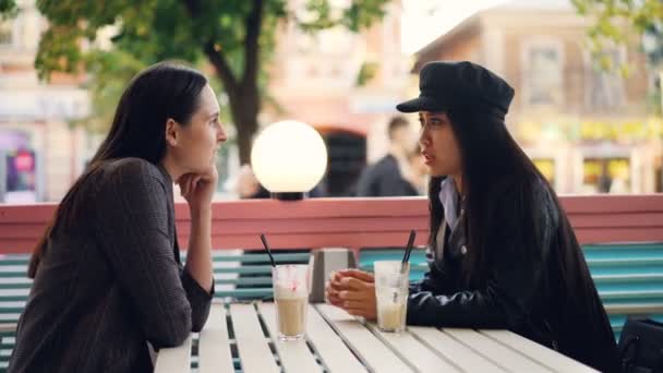 Las amigas están compartiendo noticias hablando sentadas en la mesa en una cafetería al aire libre en un cálido día de otoño y tomando cócteles. Calle peatonal ocupada es visible . — Vídeo de stock