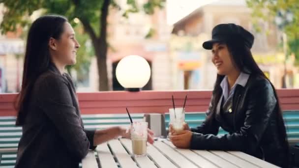 Szczęśliwy dziewcząt kaukaski i azjatyckich przyjaciele siedzą przy stoliku w kawiarni ulicy rozmawiając i sakiewkach brzęczały szklanki z koktajle korzystających z wolnego czasu. Koncepcja ludzie i napoje. — Wideo stockowe
