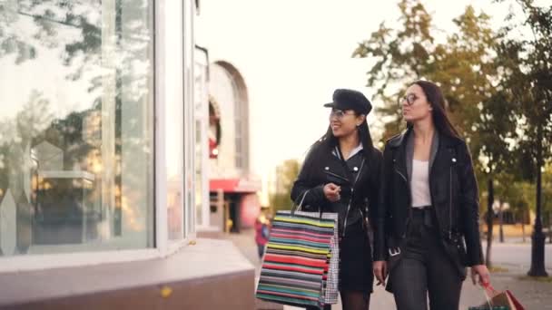 Las niñas alegres adictas a las compras están mirando los escaparates, apuntando a los bienes, hablando y riendo caminando por la calle con bolsas de papel. Estilo de vida juvenil y concepto de tiendas . — Vídeos de Stock