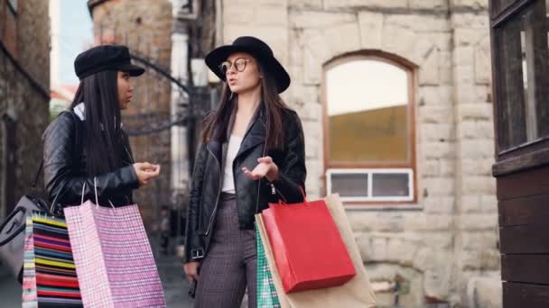 Söt asiatisk tjej pratar med sin kaukasiska vän elegant brunett stående på gatan håller kassar och skrattar. Människor, staden och shopaholics koncept. — Stockvideo