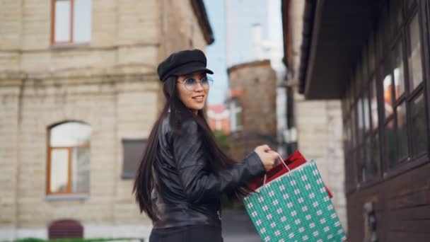 Retrato en cámara lenta de una mujer asiática sonriente adicta a las compras caminando por la calle con bolsas de papel, girando y mirando a la cámara disfrutando de las compras y la ciudad . — Vídeos de Stock