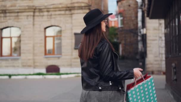 Retrato em câmera lenta de bela morena andando ao longo da rua com sacos de compras, em seguida, se voltando para a câmera e sorrindo olhando para a câmera. Conceito de juventude e consumismo . — Vídeo de Stock