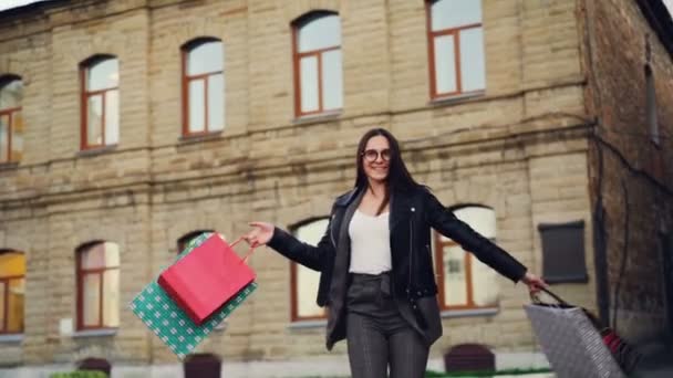 Mouvement lent de la jeune femme heureuse shopaholic dansant dans la rue avec des sacs à provisions tournant tournant regardant la caméra. Style de vie urbain et concept de consumérisme . — Video