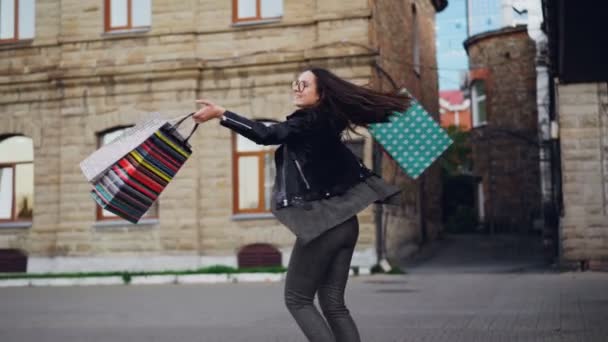 Zeitlupenporträt eines fröhlichen Mädchens, das im Freien tanzt, Einkaufstüten dreht und in die Kamera blickt. Frau trägt trendige Kleidung und Brille. — Stockvideo