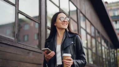 Ağır çekim yalnız gitmek kahve tutarak ve smartphone kullanarak modern şehirde yürüyüş uzun siyah saçlı güzel genç bayan. Millennials, teknoloji ve kent kavramı.
