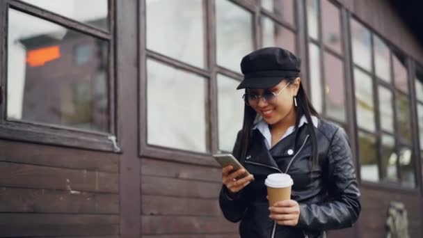 Ağır çekim gitmek içki ve gülümseyen ve ekran dokunaklı Gadget'ı kullanma smartphone tutan sokakta yürürken çekici genç kadın. Teknoloji ve duygular kavramı. — Stok video