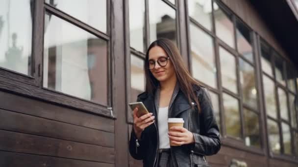 慢动作的可爱的年轻女子在眼镜使用智能手机和微笑着走在街上, 拿出咖啡。青年生活方式与城市理念. — 图库视频影像