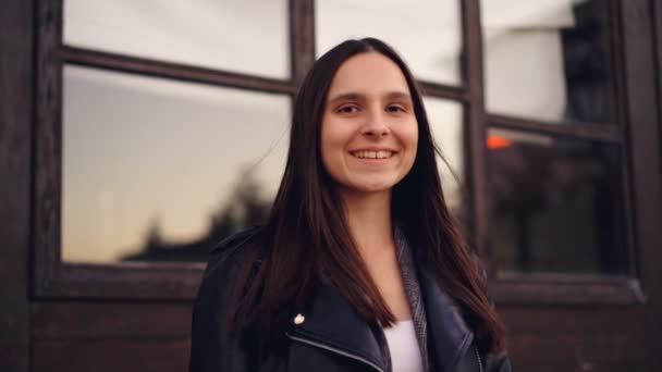 Portrait au ralenti d'une jolie jeune femme aux longs cheveux foncés et aux yeux marron portant une veste en cuir et un haut regardant la caméra et souriant. Concept de personnes, de ville et d'émotions . — Video